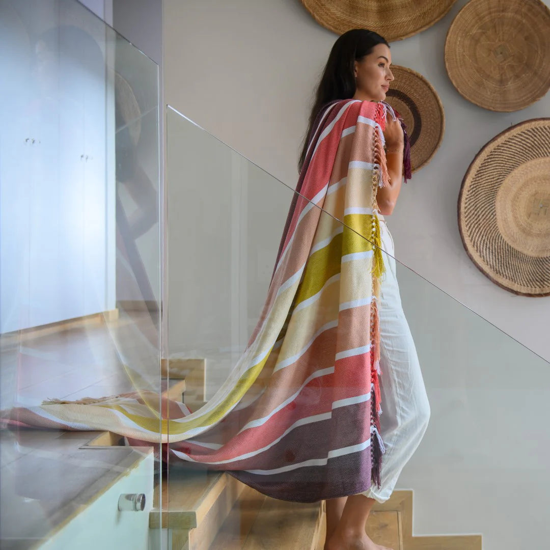 Woman standing on a staircase wearing a Protea Pinstripe Sunset throw in Burgundy/Garnet – rich striped design with hand-knotted fringes.