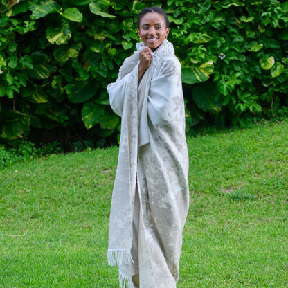 Woman wrapped Bella Vita Dappled Leaves jacquard throwstanding in a grassy area with greenery.