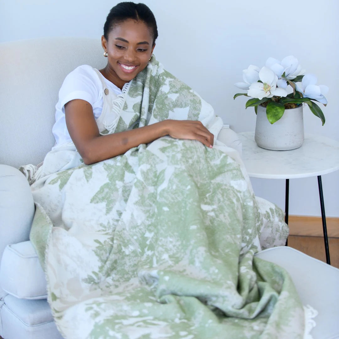 Woman wrapped in a Bella Vita Dappled Leaves jacquard throw patterned blanket sitting on a couch with a plant in the background.